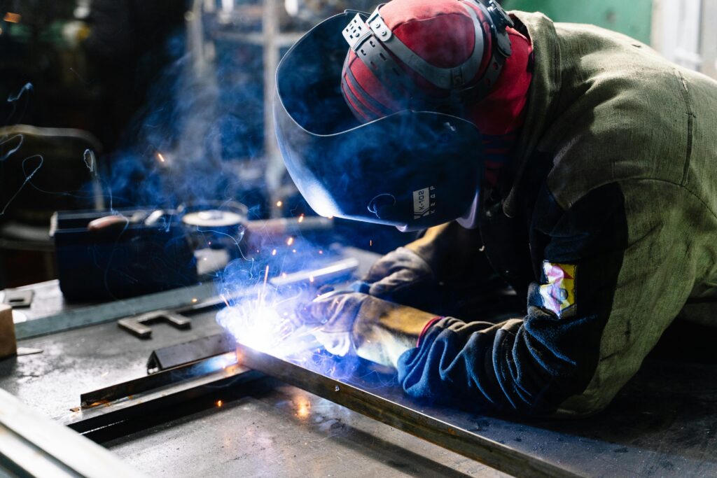 A welder works on metal fabrication indoors, creating bright sparks and smoke.